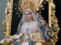 Representación de la Virgen María en un trono ornamental, vestida con una túnica blanca y un manto azul, coronada con una tiara dorada.