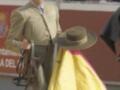 Un torero en traje tradicional sostiene un capote y una muleta, preparándose para la corrida de toros. La imagen muestra un ambiente festivo con espectadores en el fondo, enfocándose en la figura del torero.