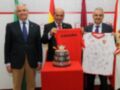 Tres hombres en trajes posan con una camiseta firmada y un trofeo de la Copa del Rey en el fondo.