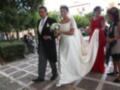 Una pareja casada camina por una calle con un vestido blanco y un traje negro. La novia lleva un velo largo y una corona de flores, mientras el novio sostiene su mano. A la derecha, una mujer en rojo y otra en negro observan el momento con alegría. El fondo muestra un edificio con balcones y árboles verdes.