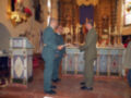 Imagen de dos hombres en uniforme militar realizando una ceremonia religiosa en un altar decorado con velas y flores.