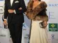 Elegante pareja en un evento formal, con el hombre en traje negro y la mujer en vestido largo beige con abrigo de pelaje. Fondo con logos de 'la Caixa' y 'EUSA'.