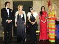 Una escena formal con cinco personas vestidas de manera distintiva. A la izquierda, un hombre en traje negro y corbata negra mira a una mujer con vestido largo azul oscuro. A la derecha, dos mujeres en vestidos largos de colores vibrantes y una mujer con un disfraz de gato. La escena parece ser parte de una fiesta o evento formal.