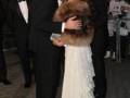 Una pareja en trajes de gala se besa mientras otros asistentes observan. La imagen muestra una celebración formal con vestimentas elegantes y un ambiente festivo.