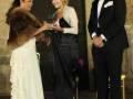 Una mujer en un vestido blanco con flecos y una peluca marrón, sostiene un trofeo mientras sonríe. Una mujer con una peluca rubia y vestido negro sostiene el trofeo, mientras un hombre en traje negro y corbata sostiene la mano de la mujer con peluca rubia.