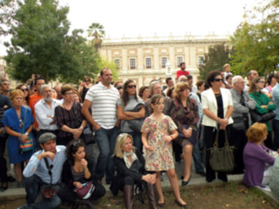 Una multitud de personas se reúne en un parque, frente a una construcción histórica. La gente está sentada y de pie, algunas con sombrillas, otros con bolsos. La escena es tranquila y parece ser un evento público o una reunión de grupo.