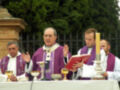 Un sacerdote en vestimenta morada y blanco, con un libro rojo, gestiona una ceremonia religiosa al aire libre. Detrás de él, otros sacerdotes y un altar con copas y velas.