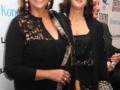 Dos mujeres en un evento formal, posando frente a una pantalla con logotipos de teatros y empresas.