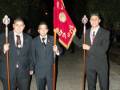Cuatro jóvenes en trajes formales sostienen una bandera roja con símbolos dorados y varas ceremoniales, posando al aire libre.