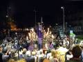 Una procesión nocturna en una ciudad, con una figura vestida de morado y un grupo de personas vestidas de blanco rodeándola. La iluminación es tenue, destacando la figura central y las luces de fondo.