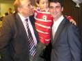 Dos hombres en trajes posan con un niño en medio, uno de ellos sostiene a un niño vestido con una camiseta roja y blanca. La escena parece ser en un evento formal, con una pantalla de fondo que muestra a varias personas.
