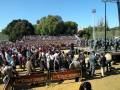 Una multitud asiste a un evento al aire libre con stands de Cruzcampo en el fondo. La escena está rodeada por árboles y cielo azul, con un público que parece estar disfrutando del evento.