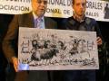 Dos hombres sostienen un cuadro que representa una escena de corrida de toros. El fondo muestra un cartel con texto en español.