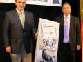 Dos hombres en trajes sostienen un cartel con ilustraciones de toros y texto. El fondo muestra una pancarta que dice "NACIONAL DE MAYORALES".