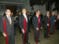 Un grupo de hombres en trajes formales, algunos sujetando bastones rojos. La escena parece ser una celebración o ceremonia formal, con luces y decoraciones en el fondo.