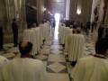 Una procesión religiosa en una iglesia con monjes vestidos de blanco y una luz brillante en el fondo.