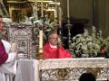 Un sacerdote en un hábito rojo se encuentra frente a una mesa de altar decorada con flores y velas, en un ambiente religioso.