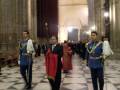 Guardias de honor en uniforme azul y rojo, caminando por un pasillo con columnas altas.