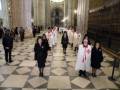 Una imagen de una ceremonia religiosa en un gran edificio con columnas y arcos, donde personas vestidas formalmente caminan por el suelo de azulejos. Algunas están con vestidos blancos y otros con trajes oscuros, mientras que una figura central lleva un hábito blanco y un cíngulo rojo. La escena sugiere una celebración o evento importante dentro de un lugar histórico y monumental.