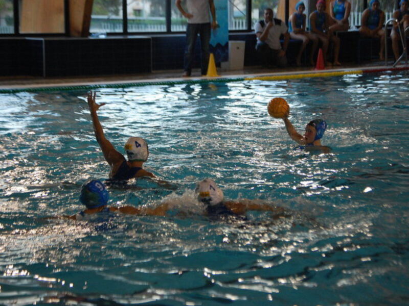 Jugadores de waterpolo en acción durante un partido en una piscina interior, con un árbitro observando desde el borde del agua.
