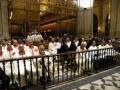 Una procesión religiosa en una catedral, con sacerdotes vestidos de blanco y rojo, frente a un altar dorado.