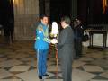 Un hombre vestido con un traje azul y una chaqueta dorada, sosteniendo un instrumento musical, mientras otro hombre en traje negro lo observa. Ambos están en una sala con suelo de azulejos y estructuras de madera en el fondo.