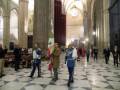 Imagen de un interior de una catedral con arcos altos y columnas. Personas vestidas formalmente, incluyendo uniformes militares, caminan por el pasillo. En la parte superior se puede ver un reloj en la pared y una bandera ondeando.