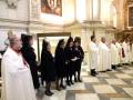 Una ceremonia religiosa en un templo, con varios personajes vestidos de negro y otros en trajes ceremoniales blancos. La imagen muestra a un grupo de personas, incluyendo una persona en el centro que parece ser la protagonista de la ceremonia. La decoración del interior incluye elementos arquitectónicos y pinturas en el techo.