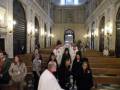 Una iglesia con una multitud de personas sentadas en las bancas. La imagen muestra a varias personas vestidas con trajes formales, algunas de ellas son sacerdotes. La decoración interior es elegante con detalles arquitectónicos y una gran puerta de madera.
