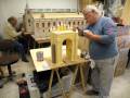 Dos hombres trabajando en un modelo de arco de piedra, con pinturas y herramientas alrededor.