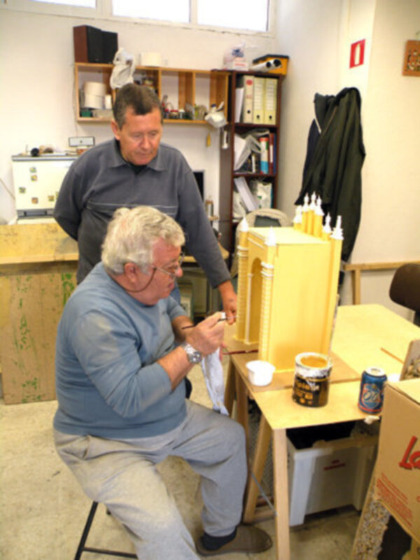 Un hombre joven observa atentamente a un anciano sentado en una silla, ambos están en una oficina con estantes llenos de libros y archivos. En la mesa hay un recipiente de café, una taza blanca vacía y un bloc de notas. La oficina tiene un ambiente tranquilo con una computadora y otros objetos de oficina en el fondo.