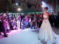 Una modelo posa en un escenario iluminado, vestida con un vestido blanco largo y elegante. La escena está llena de fotógrafos y espectadores, con luces y cámaras en el fondo.