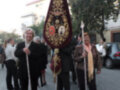 A una procesión con personas portando un escudo y una bandera. La gente está vestida formalmente, lo que sugiere que es un evento importante o festivo. La escena se desarrolla en una calle con árboles y edificios al fondo.