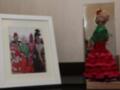 Una escena festiva con personas vestidas en trajes tradicionales, posiblemente de España. A la derecha, una muñeca vestida en un traje rojo y verde esculpido con detalles elaborados. La escena combina artefactos tradicionales y una muñeca decorativa, destacando la cultura y el arte.