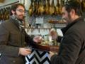 Dos hombres conversan en un ambiente con jamones colgados y una decoración de azulejos negros y blancos.