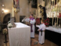 En una iglesia, un sacerdote realiza una ceremonia religiosa mientras un niño pequeño observa. La imagen muestra detalles de la decoración, incluyendo flores y velas encendidas.