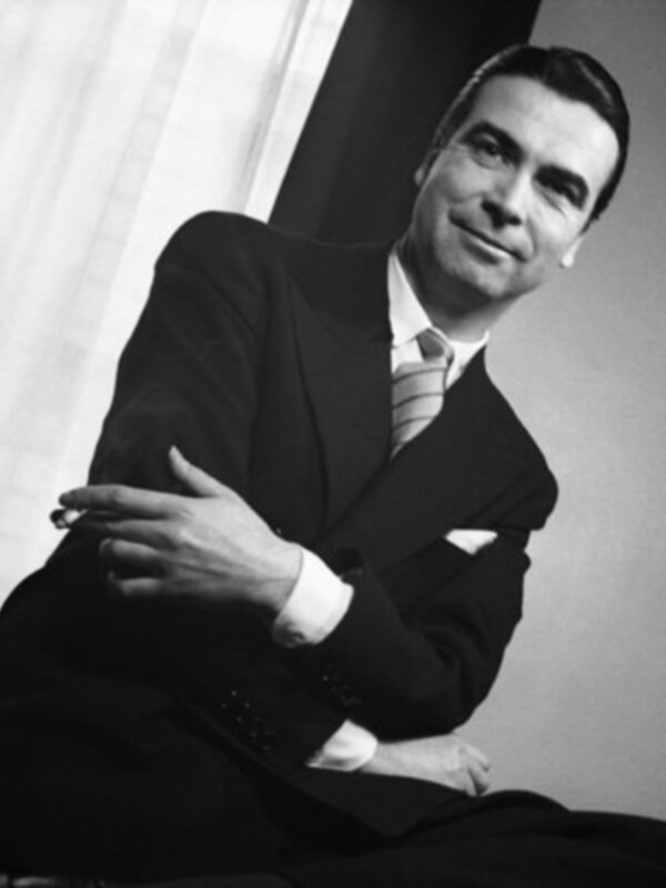 Un hombre vestido con un traje negro y una camisa blanca, posando con una sonrisa en la cara.