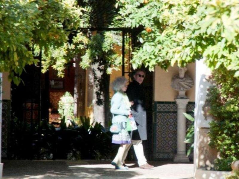 Dos personas caminan por un camino rodeado de árboles frondosos. La imagen muestra una escultura en un pedestal a la derecha y una puerta adornada con azulejos de color verde. La atmósfera es tranquila y soleada, típica de un entorno urbano con elementos históricos.