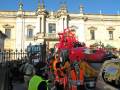 Una caravana con un personaje rojo grande en la parte delantera, frente a una iglesia histórica con detalles barrocos. Personas en uniformes de trabajo y un policía en el fondo.