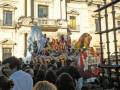 Una procesión religiosa en un ambiente histórico, con una imagen de la Virgen y figuras religiosas en el centro, rodeada por un gran público. La imagen se toma frente a una fachada de estilo clásico, con detalles arquitectónicos.