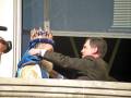 Un hombre en traje negro ajusta la corona de un hombre vestido con una túnica dorada y azul. La escena se realiza desde un balcón con una ventana de fondo, iluminada por el sol.