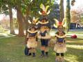 Tres individuos vestidos con trajes tradicionales de piel y plumas, posando en un parque con árboles y hojas de otoño.