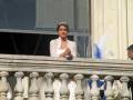 Una mujer con un vestido blanco y tocado de plumas azules, posiblemente una princesa, mirando desde un balcón.