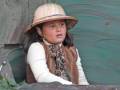 Niña con sombrero de pescador y chaleco de piel, sentada en un entorno natural, posiblemente en una excursión o viaje.