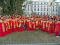 Una agrupación de personas vestidas con trajes tradicionales rojos y dorados, posando frente a un edificio blanco. Algunas personas llevan sombreros rojos y dorados, mientras que otras tienen gorros de color plateado. La imagen se toma en un entorno verde con árboles y una estructura metálica visible al fondo.