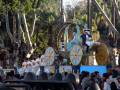 Un carro de fantasía con un personaje vestido como un príncipe azul en el medio, rodeado de decoraciones doradas y flores. El carro está flanqueado por figuras vestidas con trajes blancos y sombreros, con un fondo de árboles verdes.