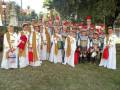 Un grupo de niños vestidos con trajes medievales y armaduras posa para una foto en un parque. Los niños llevan sombreros y vestimentas coloridas, con algunos portando bolsos rojos. La escena está rodeada de árboles y una casa en el fondo.
