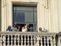 Un grupo de personas, incluyendo a un hombre con una corona en la mano, se encuentran en un balcón de una edificación histórica. La imagen muestra a varios individuos vestidos formalmente, posiblemente celebrando o participando en un evento importante. La escena transmite una sensación de solemnidad y celebración, con la presencia de la corona añadiendo un toque ceremonial.
