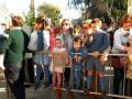Una multitud de personas, incluyendo niños y adultos, esperan en una fila detrás de un parapeto. La imagen muestra a varias personas con ropa casual, algunas sosteniendo bolsas y otros objetos. La escena parece ser una fila de espera para un evento o evento público, con árboles y edificios al fondo.
