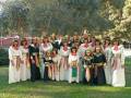 Una familia de 15 personas vestidas en trajes tradicionales, posando juntos en un parque con árboles y una estructura de metal al fondo.
